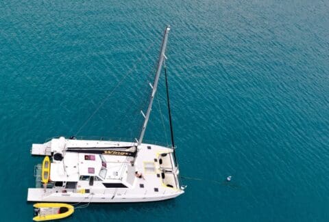 Aerial view of a sailboat named "Wings" anchored in the clear blue waters of the Whitsundays. A small yellow dinghy floats beside it, epitomizing the serene experience offered by Wings Sailing Charters.