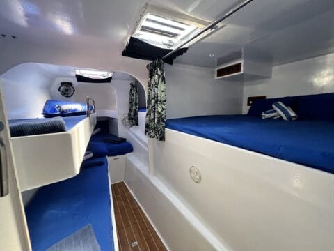 The interior of the compact boat cabin, ideal for Whitsundays sailing charters, features multiple beds with blue bedding, a wooden floor, and a small window on the ceiling.